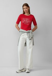 Rood T-shirt met witte grafische tekst, gecombineerd met lichtgekleurde wijde broek en een grijze sweater om de taille geknoopt. Lichte sneakers maken de look af.
