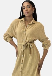 Beżowa sukienka zapinana na guziki, wykonana z teksturowanej tkaniny; posiada przewiązany w pasie wygląd, długie rękawy i luźny krój. Dyskretna kratka w całym materiale.