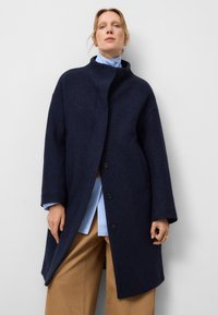 Manteau en laine bleu marine avec un col haut, une ouverture à l'avant et de grands boutons. Présente des manches larges et une coupe décontractée, associé à une chemise bleu clair.