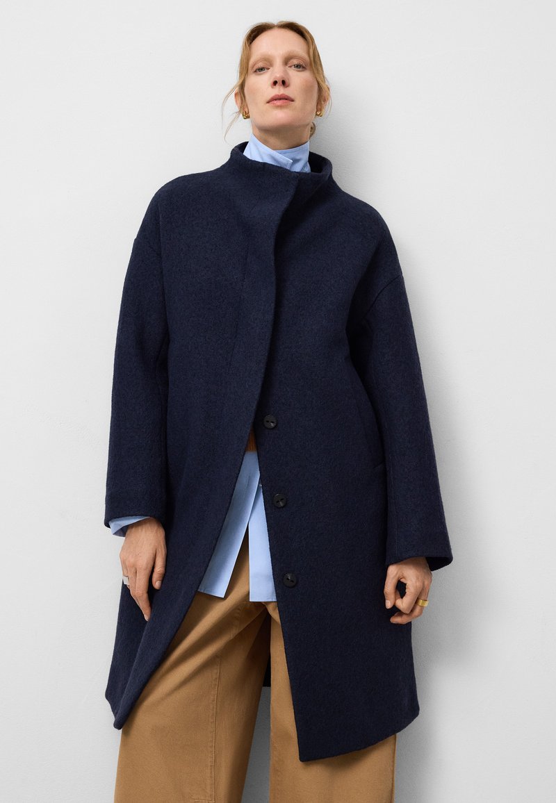 Manteau en laine bleu marine avec un col haut, une ouverture à l'avant et de grands boutons. Présente des manches larges et une coupe décontractée, associé à une chemise bleu clair.