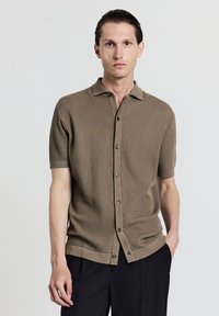 Kurzärmliges, kragenloses Strickhemd in Beige mit strukturiertem Muster, ausgestattet mit dunklen Knöpfen und geradem Saum. Getragen mit dunklen Hosen.