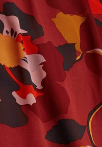 Textil med en röd bakgrund och stora blommönster i gult, orange, rosa och svart. Texturen verkar slät och mjuk.