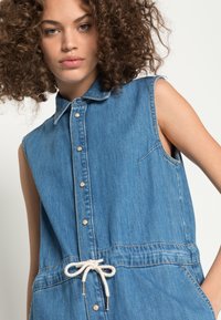 Vestido de ganga sem mangas com colarinho, fecho de botão na frente e cintura com cordão. Apresenta um bolso no lado esquerdo do peito e bordas desfiadas.