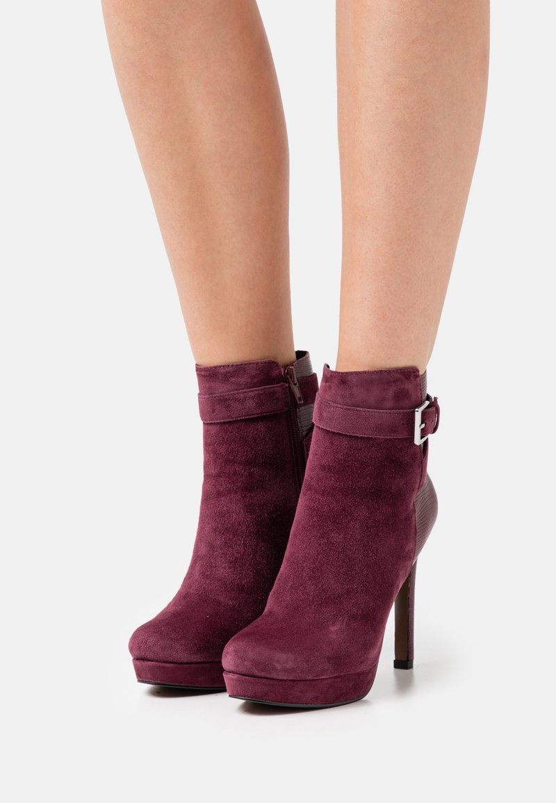 Bottes plateformes en daim bordeaux montant jusqu'à la cheville, avec fermeture éclair latérale et boucle, portées sur des jambes nues sur un fond uni.