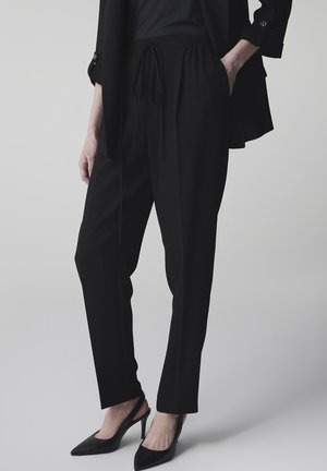 Frau trägt schwarze, maßgeschneiderte Hose mit Kordelzugbund, schwarzen Blazer und schwarze spitze Slingback-Absatzschuhe, steht mit einer Hand in der Tasche.