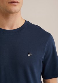 Moški v temno modri majici s kratkimi rokavi, na kateri je na levem delu prsi majhna vezenina bele ptice, prikazan od vratu do sredine trupa.