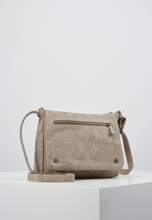 Sac bandoulière beige avec poche zippée à l'avant, rabat à deux clous métalliques, et bandoulière réglable, posé sur une surface blanche.