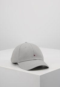 Gorra de béisbol gris claro con un pequeño logo rojo, blanco y azul en la parte frontal, colocada sobre una superficie blanca inclinada contra un fondo gris liso.
