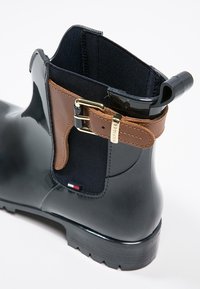 Stivale da pioggia lucido nero con un accento in pelle marrone, hardware dorato, pannello in tessuto elasticizzato e un'etichetta logo nei colori rosso, bianco e blu.