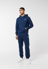 Vyras stovintis vilkintis tamsiai mėlynę gobtuvinę megztinį ir sportines kelnes su baltais sportbačiais priešais vientisą baltą foną.