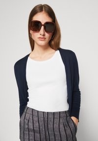 Un cardigan bleu marine foncé sur une chemise blanche, associé à un pantalon rayé gris. Le modèle porte des lunettes de soleil brunes surdimensionnées et a les cheveux bruns lisses.