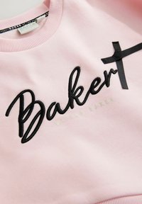 Felpa rosa con ricami neri che scrivono "Baker" e un nastro nero decorativo. Collo a coste e texture del tessuto liscia. Dettagli metallici verdi.