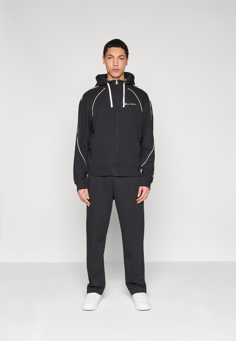 Sweat à capuche zippé noir avec accents blancs, associé à un pantalon assorti. Confectionné en tissu doux, il présente un logo sur la poitrine et une capuche à cordon.