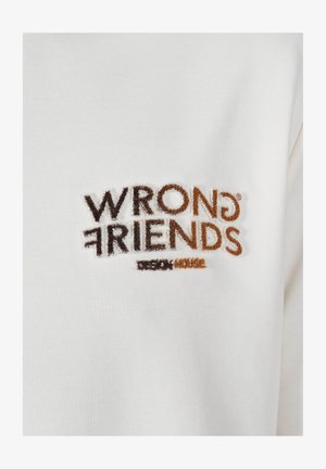 Witte getextureerde stof met een geborduurd logo in bruin en tan, met de tekst "WRONG FRIENDS" en daaronder "DESIGN HOUSE".