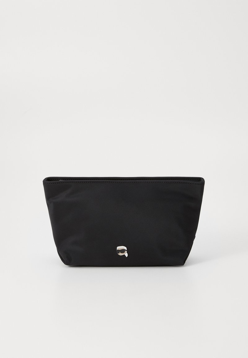 Čierna nylonová taška so zipsom na vrchu, mierne zužujúci sa dizajn; na prednej strane má minimalistický strieborný logový detail. Hladký povrch.