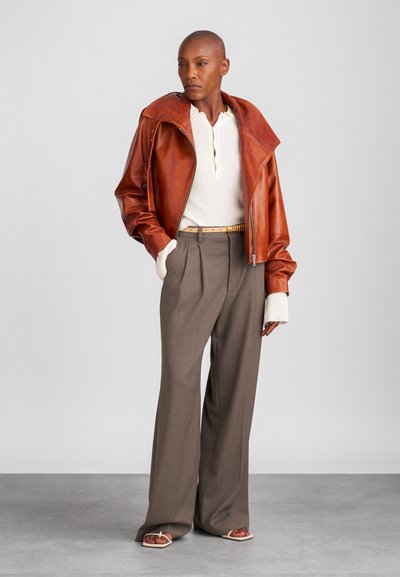 Giacca da motociclista in pelle marrone con ampio collo, henley a coste crema, pantaloni grigi a vita alta e sandali beige. Materiali testurizzati.