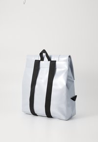 Silberner, glänzender Rucksack aus wasserdichtem Material. Verfügt über schwarze Träger, eine umklappbare Oberseite und eine Seitentasche für zusätzlichen Stauraum.