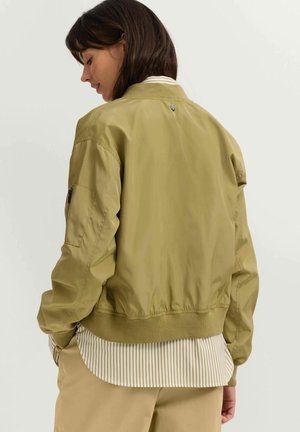 Olivgrüne Bomberjacke mit geripptem Kragen, Reißverschlusstaschen und elastischem Saum. Darüber ein gestreiftes Hemd getragen. Glattes, leichtes Material.