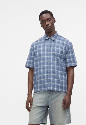 Junger Mann trägt ein blau kariertes kurzärmeliges Hemd mit Knöpfen und helle Jeansshorts mit Stern-Patches auf weißem Hintergrund.
