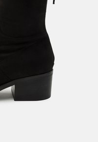 Botín de ante negro hasta el tobillo con un bajo tacón cuadrado y una textura suave, con cremallera lateral y un diseño limpio y minimalista.