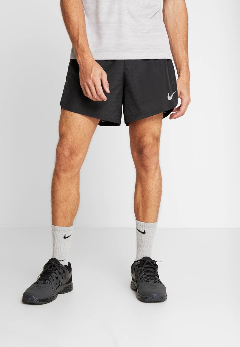 Shorts de sport noirs à texture lisse avec un accent de logo, associés à des chaussettes grises et des baskets noires, affichant un look sportif.