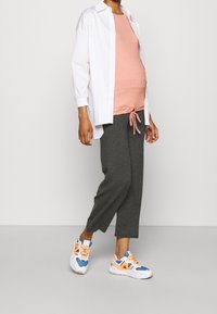 Těhotenský outfit sestávající ze světle růžového topu, volných šedých kalhot a vícebarevných tenisek. Bílá košile je rozepnutá a nošená přes vršek.