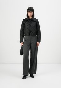 Veste courte matelassée noire avec un large col, assortie à un pantalon gris à jambes larges. Le mannequin tient un sac à main noir et porte des chaussures noires à bouts pointus.