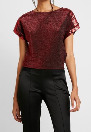 Kobieta ubrana w czerwony, cekinowy top z krótkim rękawem, w połączeniu z czarnymi, satynowymi spodniami z wysokim stanem, na jednolitym tle.