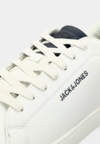 Bijela kožna tenisica s bež vezicama, s tamnoplavim podstavljenim okovratnikom i utisnutim logom "JACK&JONES" sa strane.