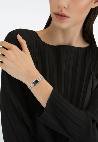 Γυναίκα με μαύρο πλισέ τοπ που φορά ασημένιο ρολόι χειρός με μαύρο εξαγωνικό καντράν, κρατώντας τον αντίθετο ώμο με το ένα χέρι.