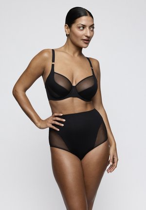 Woman posing in black mesh-accented bra and high-waisted underwear, standing with one hand on hip against plain background.