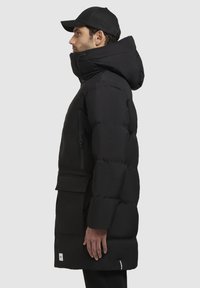 Sort pufferjakke med høj krave, lynlåslomme på siden og justerbar hætte, med en struktureret overflade og et logo-patch nær kanten.