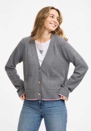 Frau lächelt und schaut zur Seite, trägt eine graue, geknöpfte Strickjacke, ein weißes Hemd mit Herzdesign und blaue Jeans.