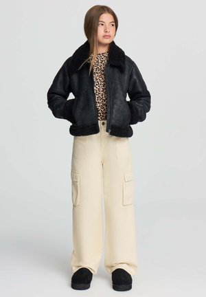 Ragazza adolescente con capelli lunghi, indossa un giacca di shearling nera, una maglietta con stampa leopardata, pantaloni cargo beige e scarpe nere, in piedi con le mani in tasca.