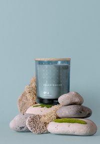 Glaskerzenbehälter mit einem Holzdeckel, der ein blaugetöntes Design aufweist. Umgeben von verschiedenen glatten Steinen und Moosakzenten.
