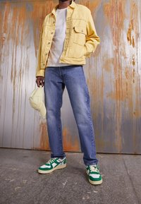 Žlutá denimová bunda, bílý tričko, modré džíny a zeleno-bílé tenisky. Drží světle žlutou čepici. Pozadí tvoří texturovaná zeď.