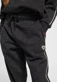 Schwarze Sweatpants aus weichem Stoff, mit einer silbernen Sprenkeltextur, elastischem Taillenbund mit Kordelzug und einem Logo auf der Hüfte.