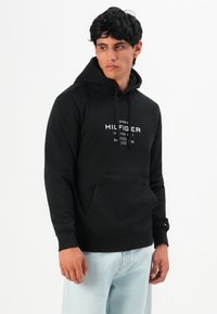 Hombre vestido con sudadera negra de Tommy Hilfiger y vaqueros azul claro, de pie contra un fondo blanco, mirando hacia un lado.