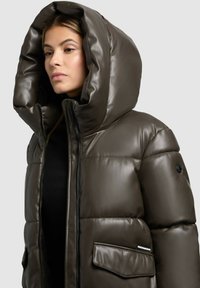 Rudas paminkštintas šalikas su dideliu, struktūrizuotu kapišonu, užtrauktuko priekyje, šoninėmis kišenėmis ir blizgiu paviršiumi; turi siuvinėtą tekstūrą ir juodą aparatūrą.