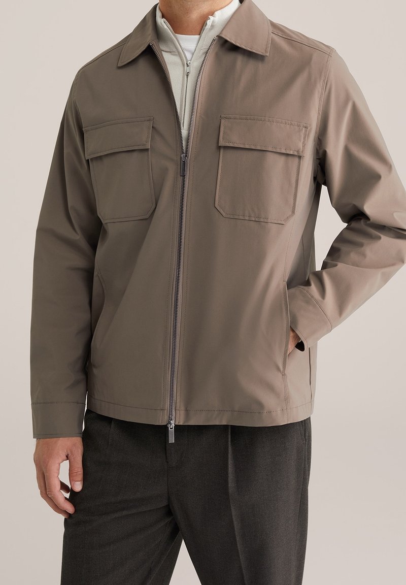 Uomo che indossa una giacca taupe con zip e due tasche a patta sul petto, pantaloni grigio scuro, con una mano nella tasca laterale, su uno sfondo neutro.