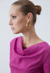 Magenta tekstil topp med en drapert cowl-hals, som har et teksturert mønster. Gull hoops øreringer fremhever stilen.
