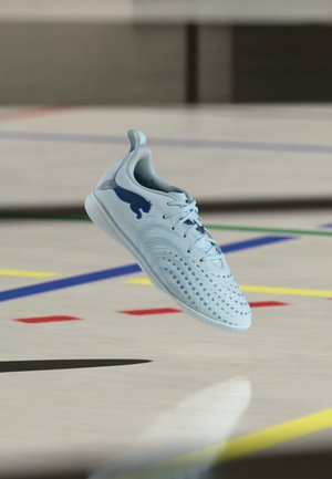 Pantof sport albastru deschis cu logo albastru închis plutind deasupra unui podea de sală marcată cu linii colorate.