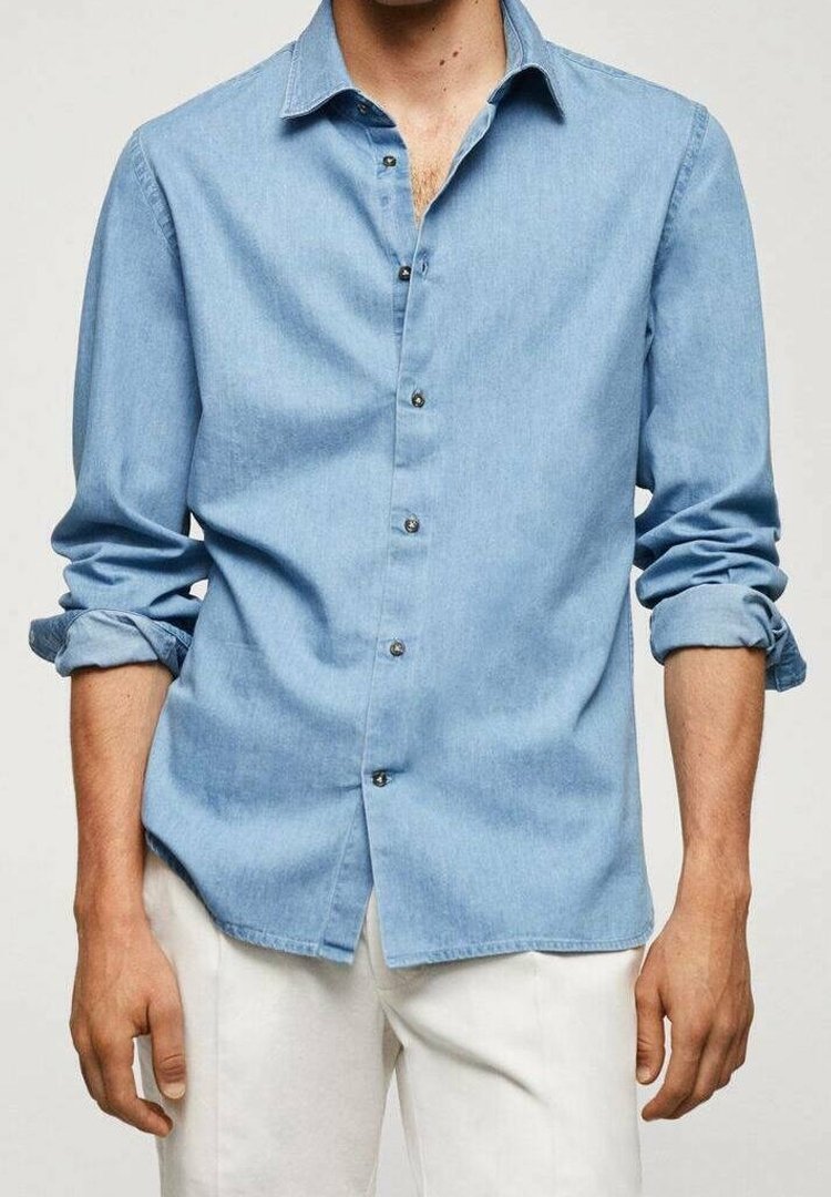 Homme portant une chemise bleu clair à boutons avec les manches retroussées et un pantalon blanc cassé, debout devant un fond uni.