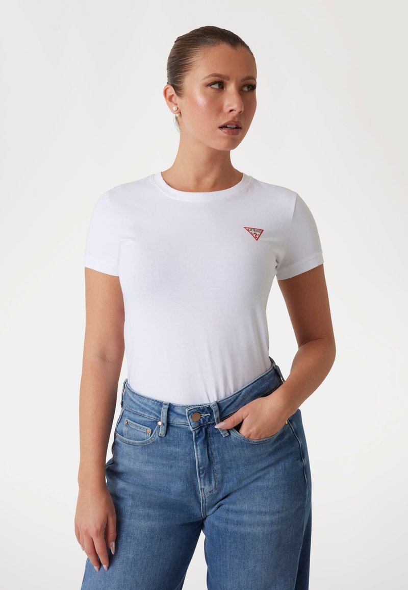 T-shirt bianca in cotone con scollatura rotonda e maniche corte, caratterizzata da un piccolo logo triangolo rosso sul lato sinistro del petto, abbinata a jeans blu chiaro.