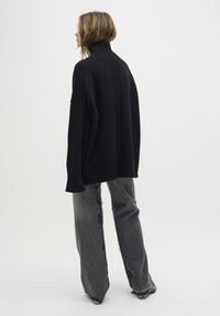 Överdimensionerad svart stickad tröja med hög ribbad krage och sänkt axel, tillsammans med slitna grå vida jeans.