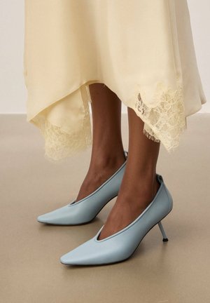 Stopy w jasnoniebieskich szpilkach z zakrzywionym noskiem, w zestawieniu z kremową spódnicą wykończoną koronką, której dół widoczny jest powyżej kostek.