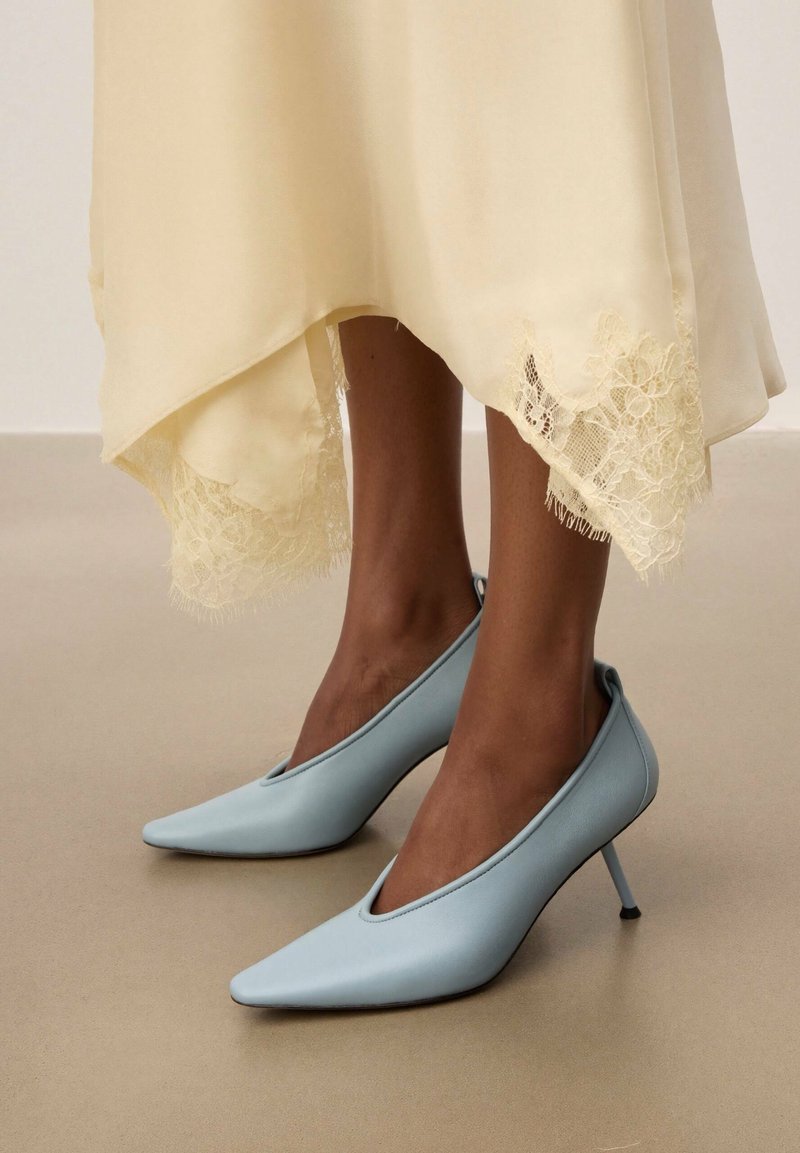 Pieds portant des escarpins à bouts pointus bleu clair, assortis à un ourlet de jupe ivoire orné de dentelle visible au-dessus des chevilles.