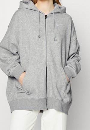 Osoba ubrana w jasnoszary rozpinany na zamek kaptur z kieszeniami i małym białym logo Nike na piersi, w połączeniu z białymi spodniami.