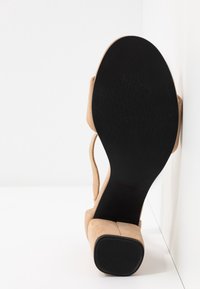 Sandale beige en daim avec semelle noire et talon bloc vue par dessous, reposant contre un angle de mur et de sol blanc.