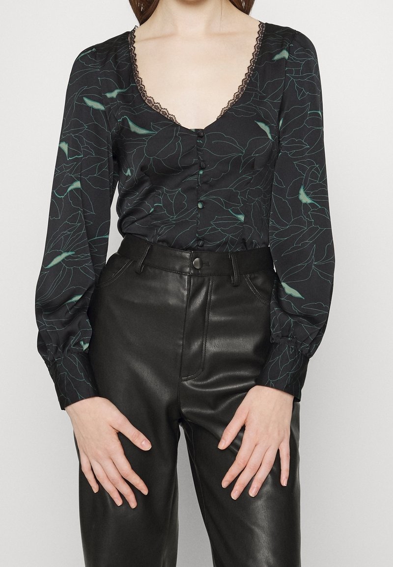 Femme portant un chemisier noir à fleurs manches longues avec un col en dentelle et un pantalon en cuir noir taille haute, les mains reposant près des cuisses.
